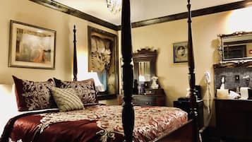 Una cama con dosel adornada con una colcha estampada en rojo y blanco, un cabecero de madera y una lámpara colgante. Se ve un aparador con espejo, un teléfono y un perchero.