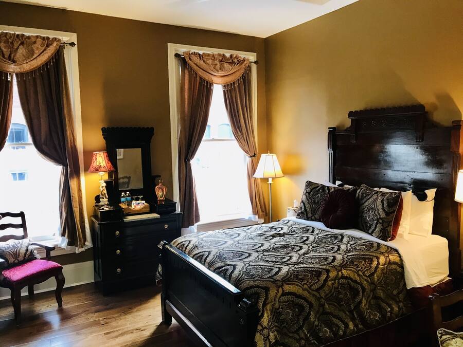 A bedroom with a large bed, a dresser, a mirror, and two windows with curtains.
