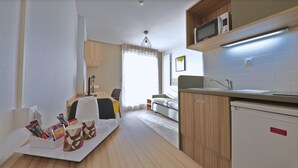 A modern kitchen with wooden cabinets, a microwave, and a refrigerator. There is a dining area with a table and chairs, and a sofa in the background. - Apparteo Dijon (Dijon)