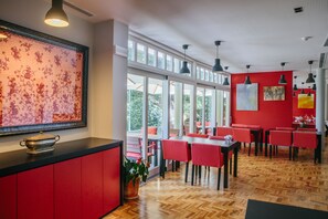 A modern dining area with red chairs, a wooden herringbone floor, and large windows offering a view of greenery.