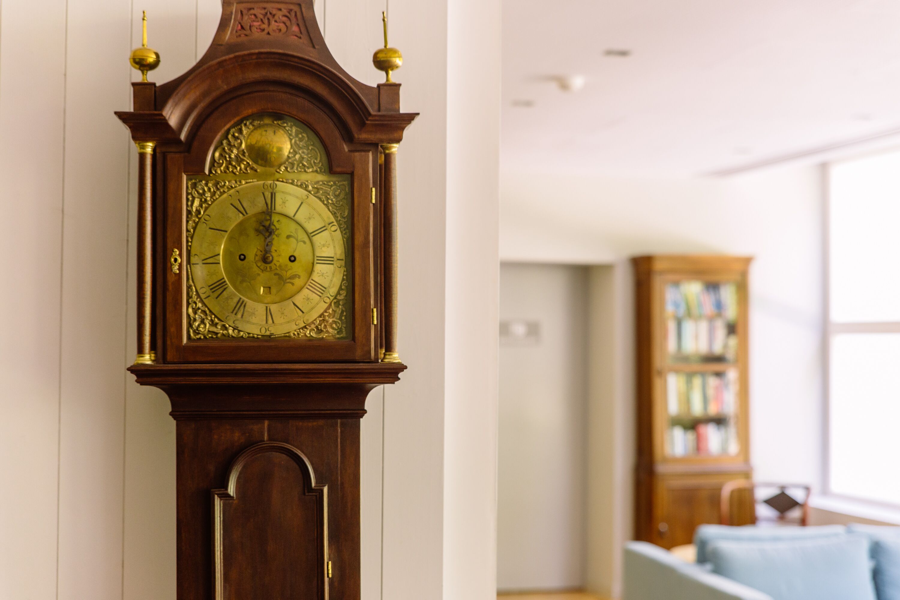 Une grande horloge comtoise richement décorée, avec un cadran doré et des chiffres romains, placée dans une pièce où l’on aperçoit une bibliothèque et une fenêtre au fond.