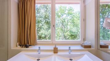 Une salle de bain avec un grand lavabo blanc, deux robinets, une fenêtre avec des rideaux bruns et une vue sur les arbres à l’extérieur.