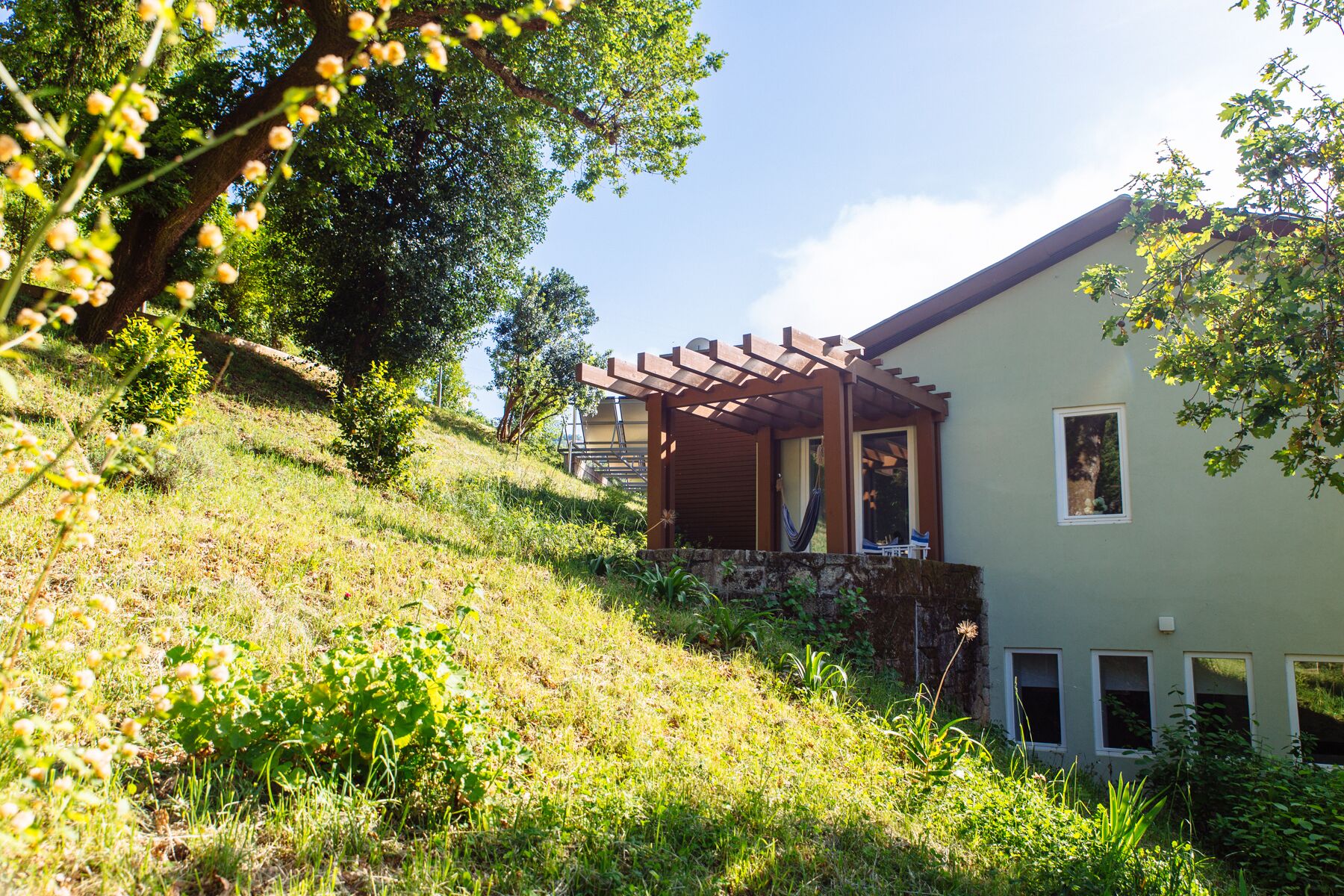 A house with a pergola on a grassy slope, surrounded by trees and vegetation.