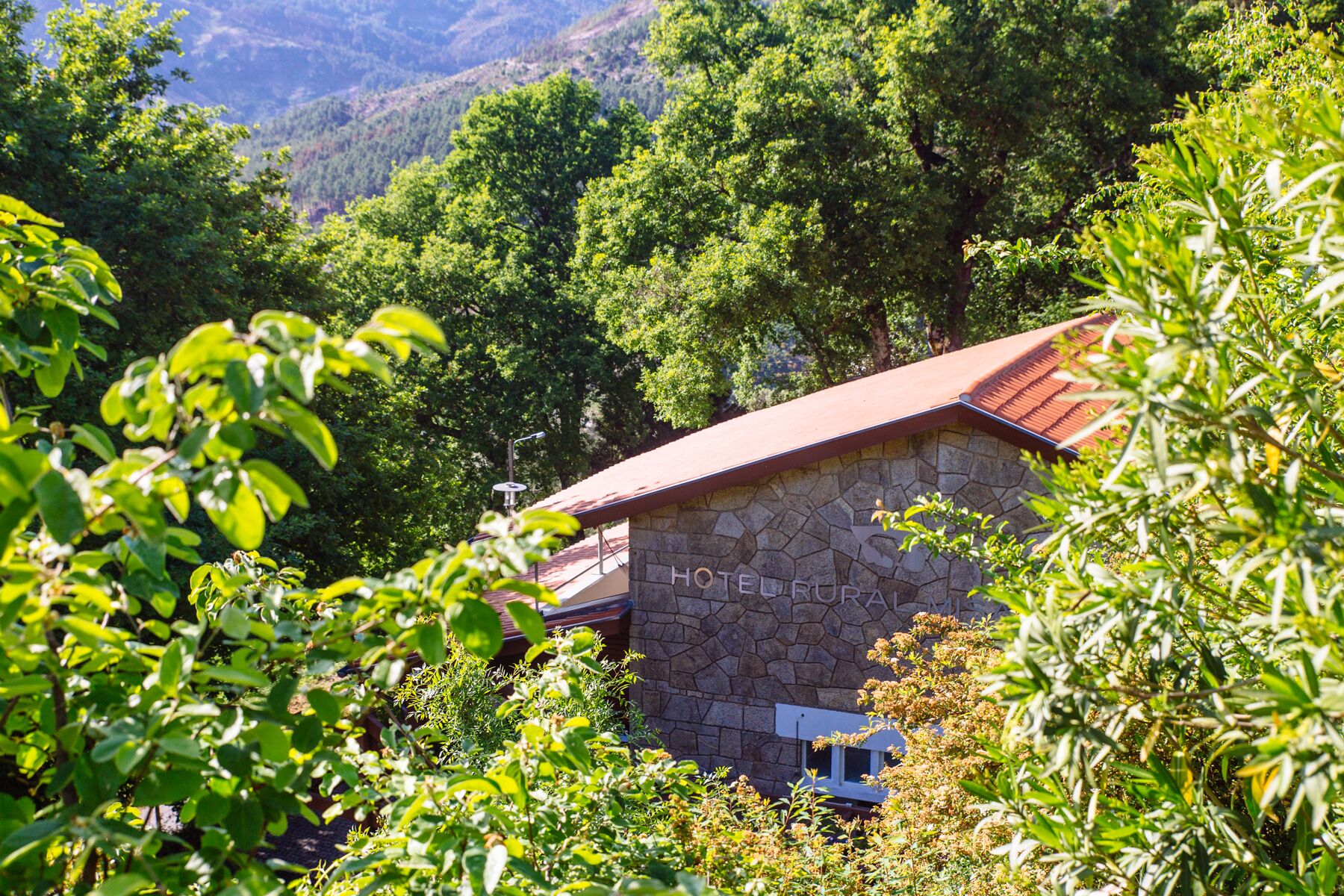 A stone building with a red roof, labeled "HOTEL RUPA," surrounded by lush greenery and mountains in the background.