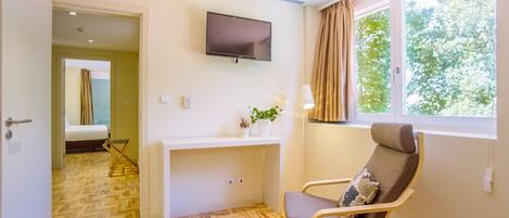 A hotel room with a wooden herringbone floor, a flat-screen TV mounted on the wall, a bedside table with a plant, a chair with a cushion, and a window with curtains.