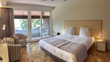 Habitación de hotel con una cama grande, una zona de estar con una silla, una mesita de noche con lámpara, y vistas al exterior a través de una puerta corredera.