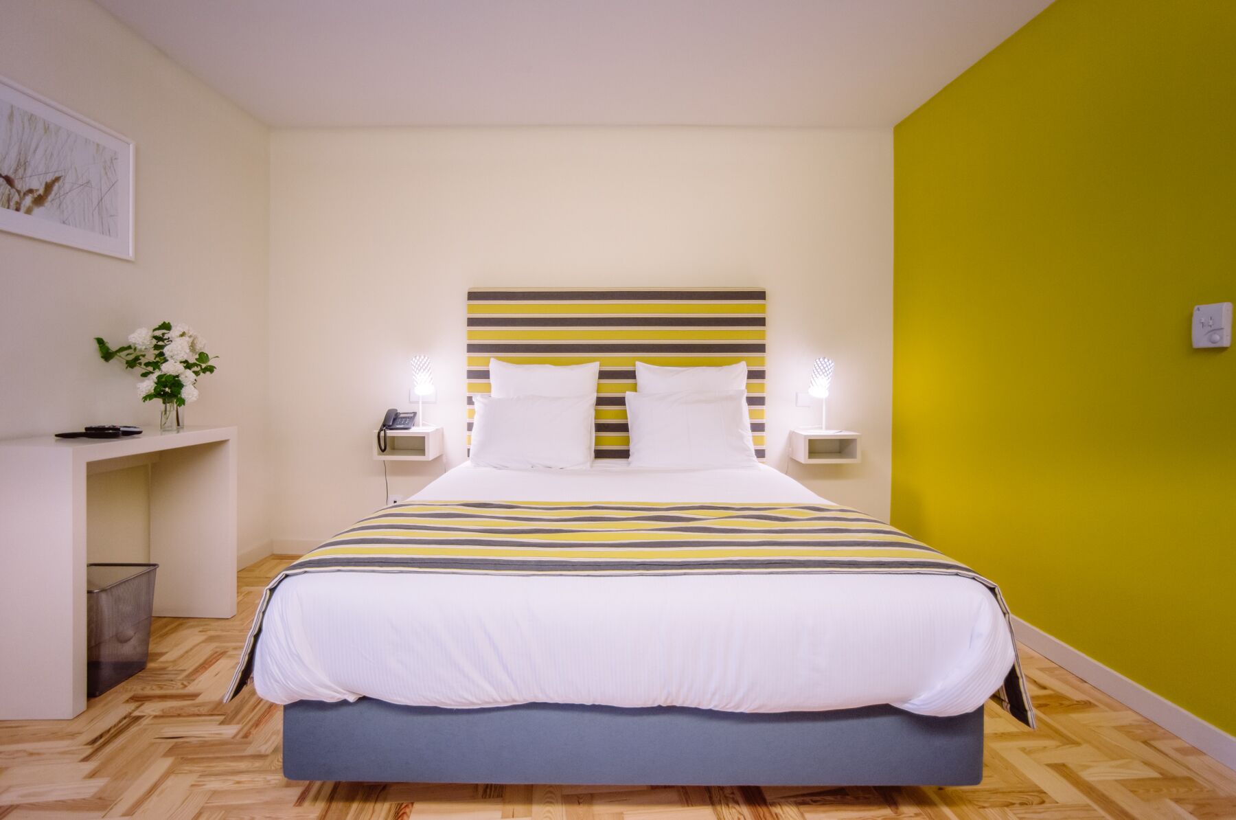 A hotel room with a bed, bedside tables, a nightstand, a lamp, and a wall-mounted light fixture.