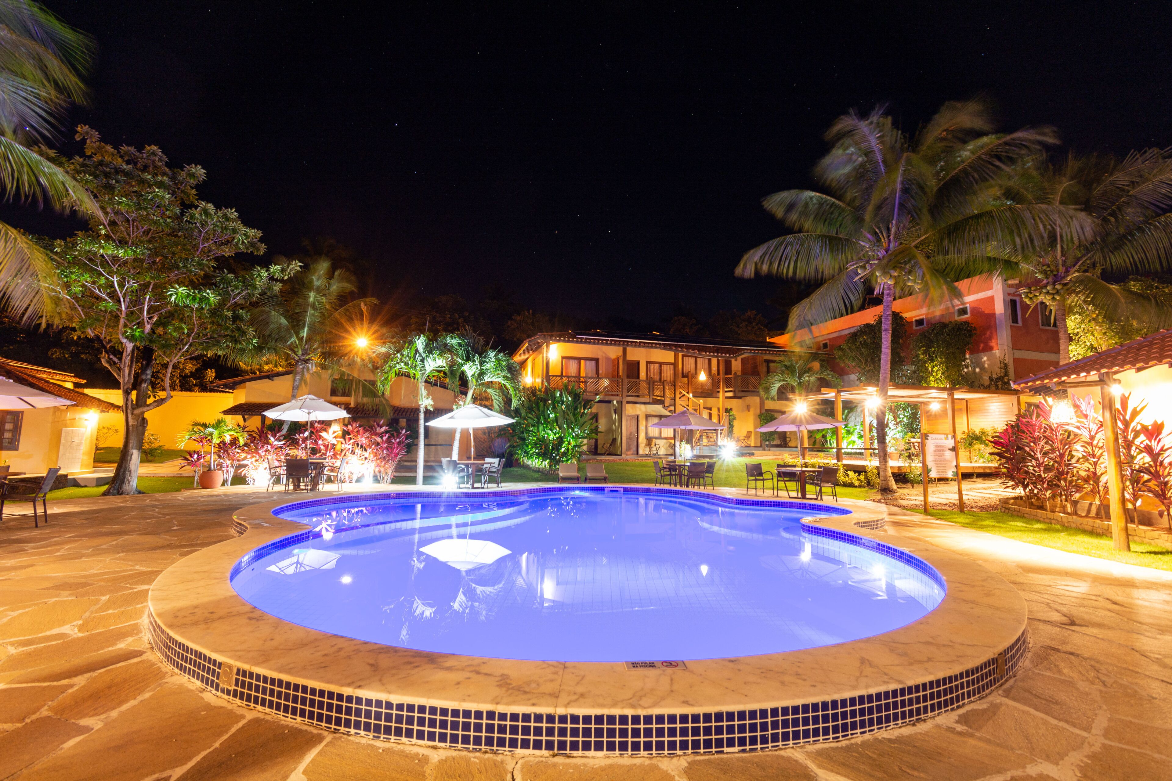 A swimming pool with a tiled edge, surrounded by stone flooring, palm trees, and outdoor seating areas with umbrellas.