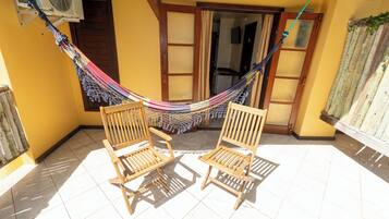 Een hangmat tussen twee houten stoelen op een betegeld terras, met een gele muur en een deur op de achtergrond.