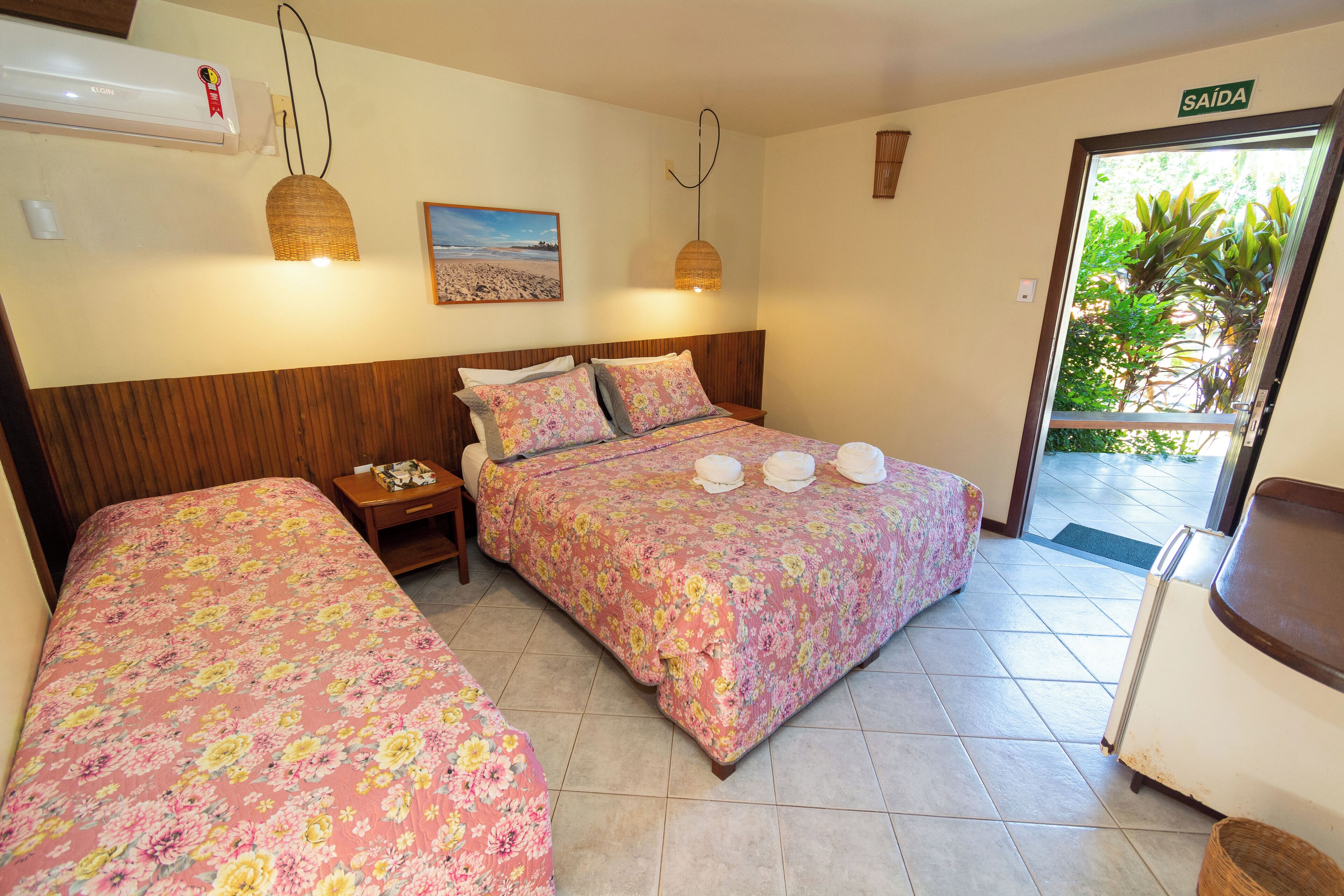 A hotel room with two beds, a nightstand, a wall-mounted air conditioner, a hanging lamp, a framed picture, and a door leading to a balcony.