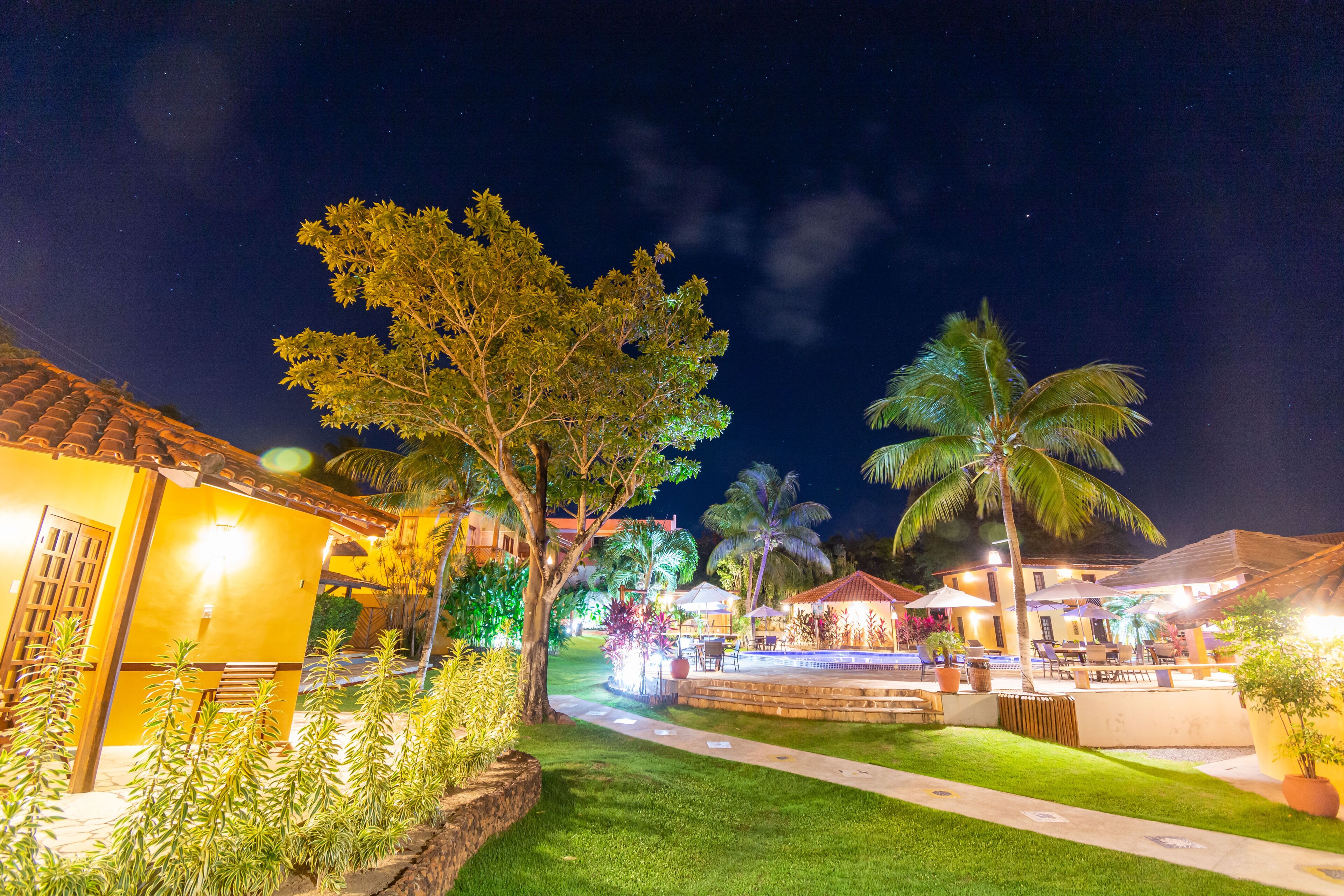 A well-lit outdoor area with a pathway, palm trees, and a building with a tiled roof.