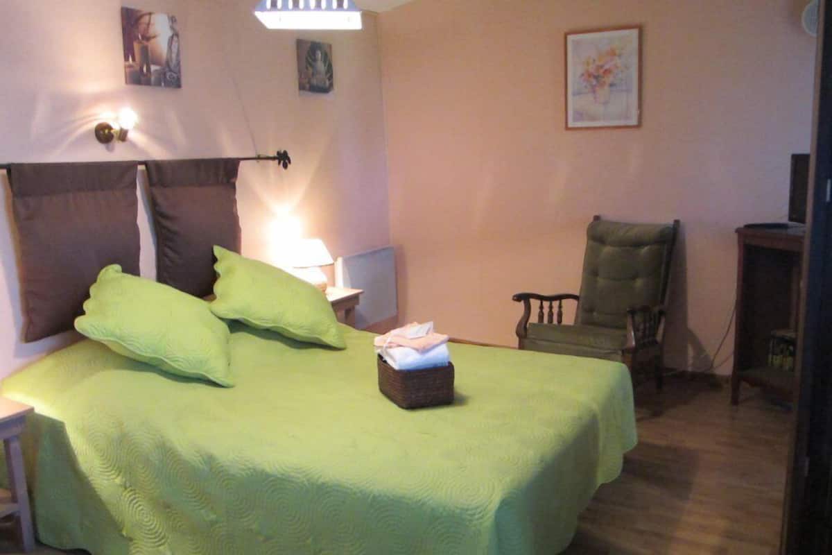 Un lit bien fait, avec une literie et des oreillers verts, une table de chevet avec une lampe, une chaise et une petite table sur laquelle se trouve un panier.