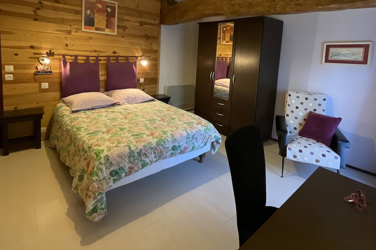 Une chambre avec un plafond en bois, un lit recouvert d’une literie à motifs floraux, une chaise agrémentée d’un coussin à pois et une armoire en bois.
