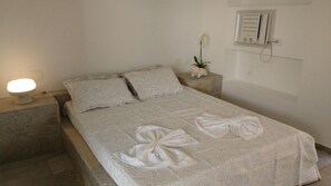 Un lit bien fait, recouvert d’une couvre-lit à motifs, une lampe de chevet, une petite plante en pot et un climatiseur fixé au mur.