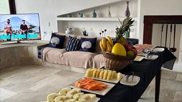 A buffet table with various fruits, cheeses, and snacks, a television displaying a travel show, and a cozy seating area with cushions.