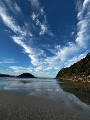 A beach with clear skies, calm water, and a forested hillside. - Pousada Pé na Areia Guarujá (Guarujá)