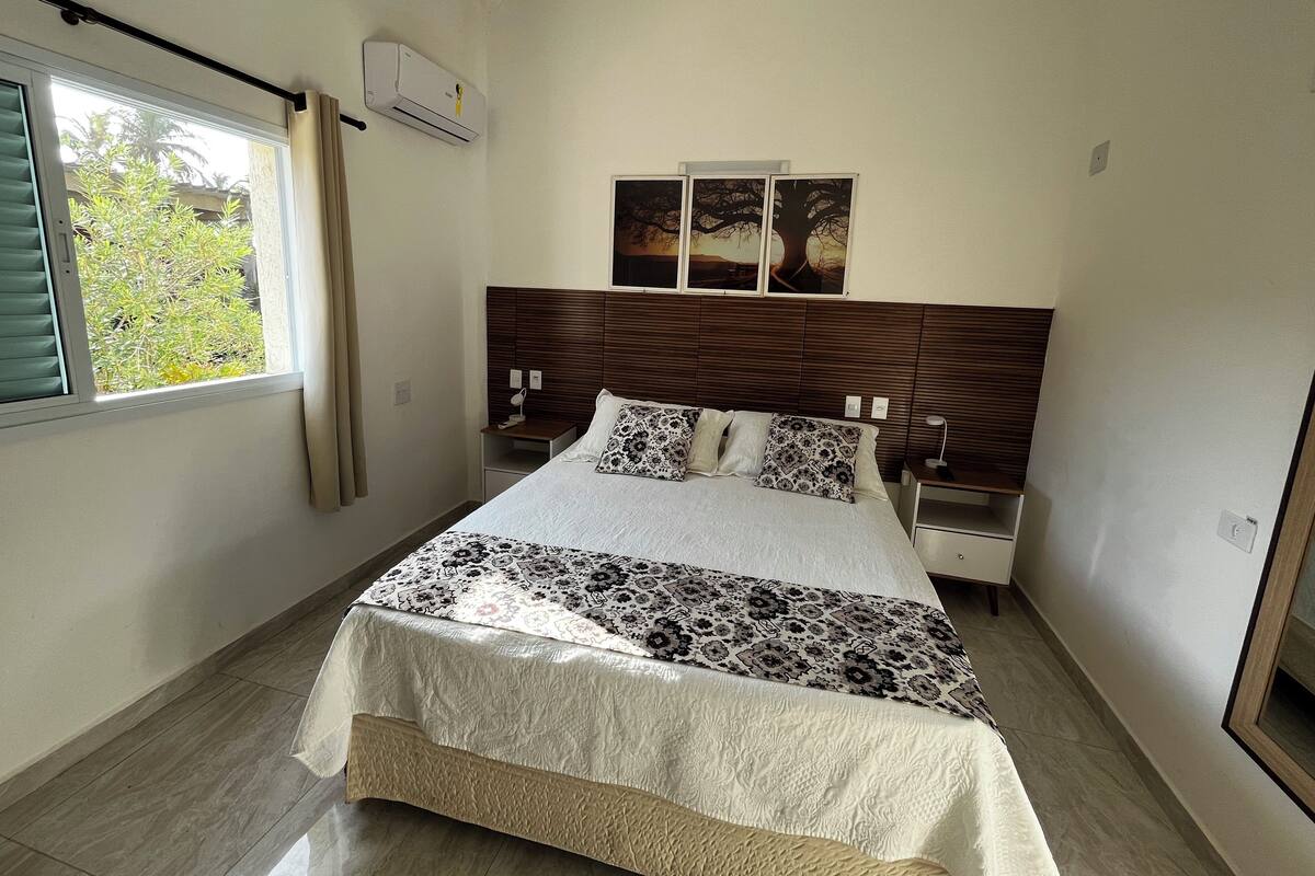 Une chambre à coucher avec un lit, une fenêtre avec des rideaux, un climatiseur, une table de chevet et une commode, ainsi qu’un tableau accroché au mur.