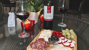 Una mesa dispuesta con una botella de vino, copas, un plato de quesos y embutidos, y un jarrón de flores.