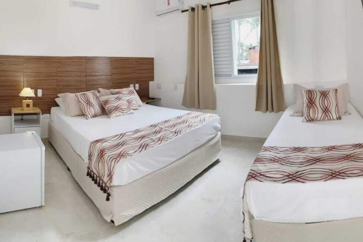 Une chambre d’hôtel avec deux lits, une tête de lit en bois, une table de chevet avec une lampe, une petite armoire blanche et un téléviseur fixé au mur.