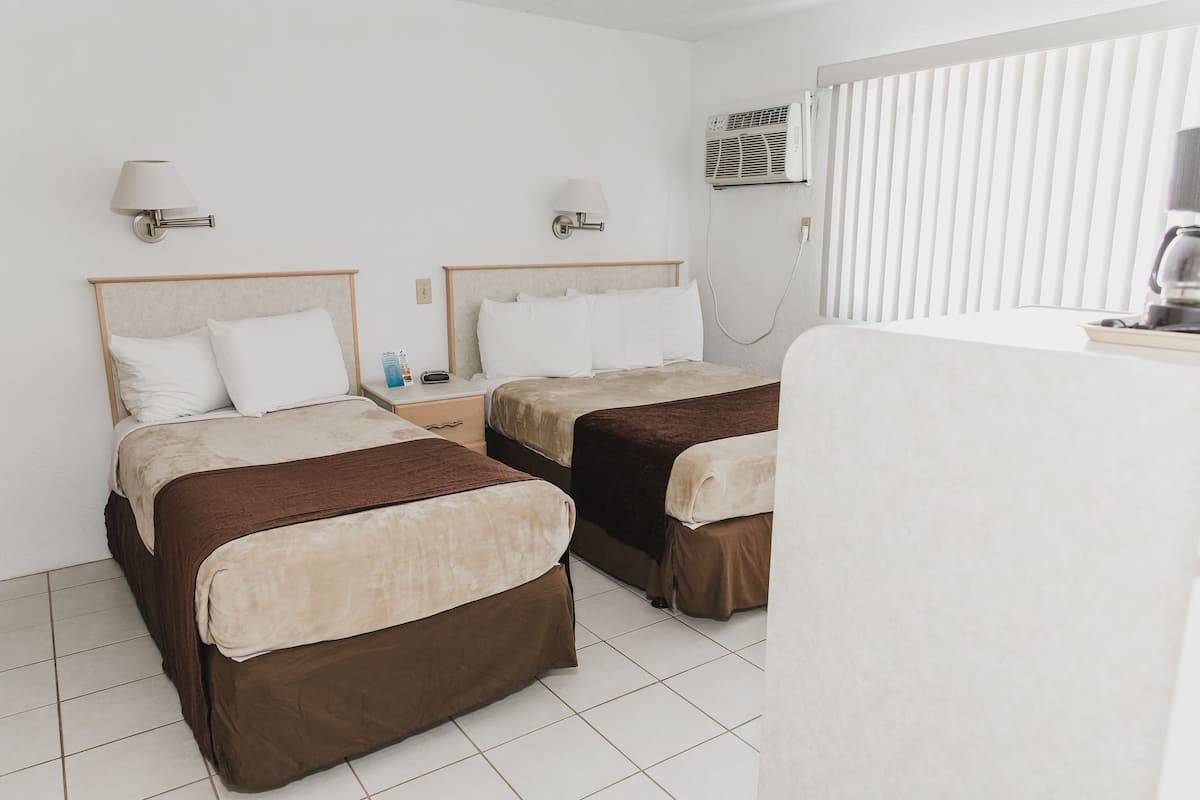 A hotel room with two beds, each with a headboard and bedside tables. There is an air conditioning unit mounted on the wall and a window with blinds.