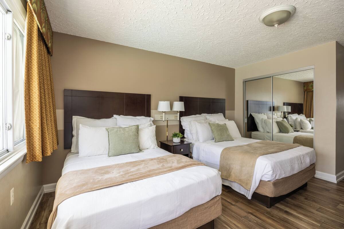 A hotel room with two beds, a large mirror, a nightstand with lamps, and a window with curtains.