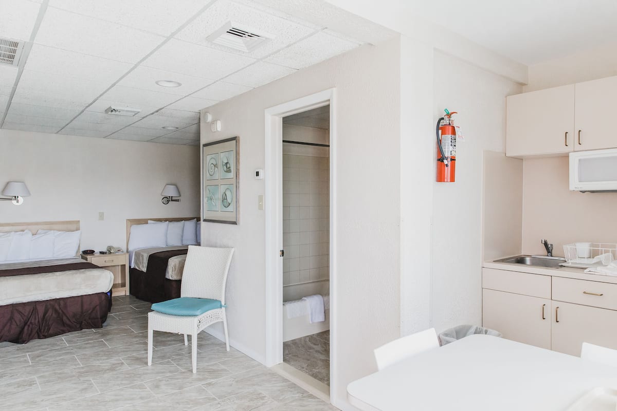 A hotel room with a bed, a chair, a small table, a kitchenette, and a bathroom with a shower.