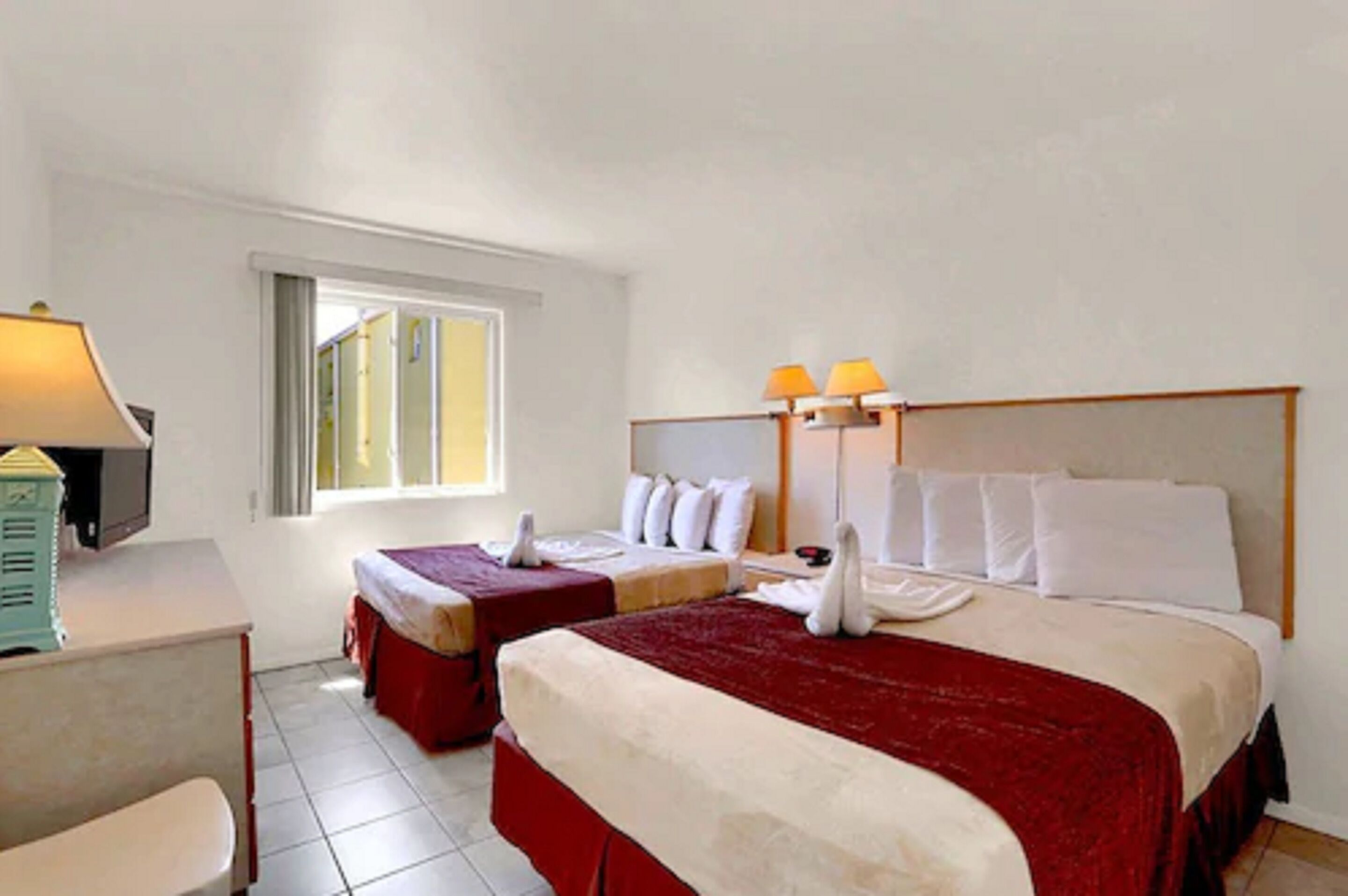 A hotel room with two beds, a desk with a lamp, and a window providing natural light.