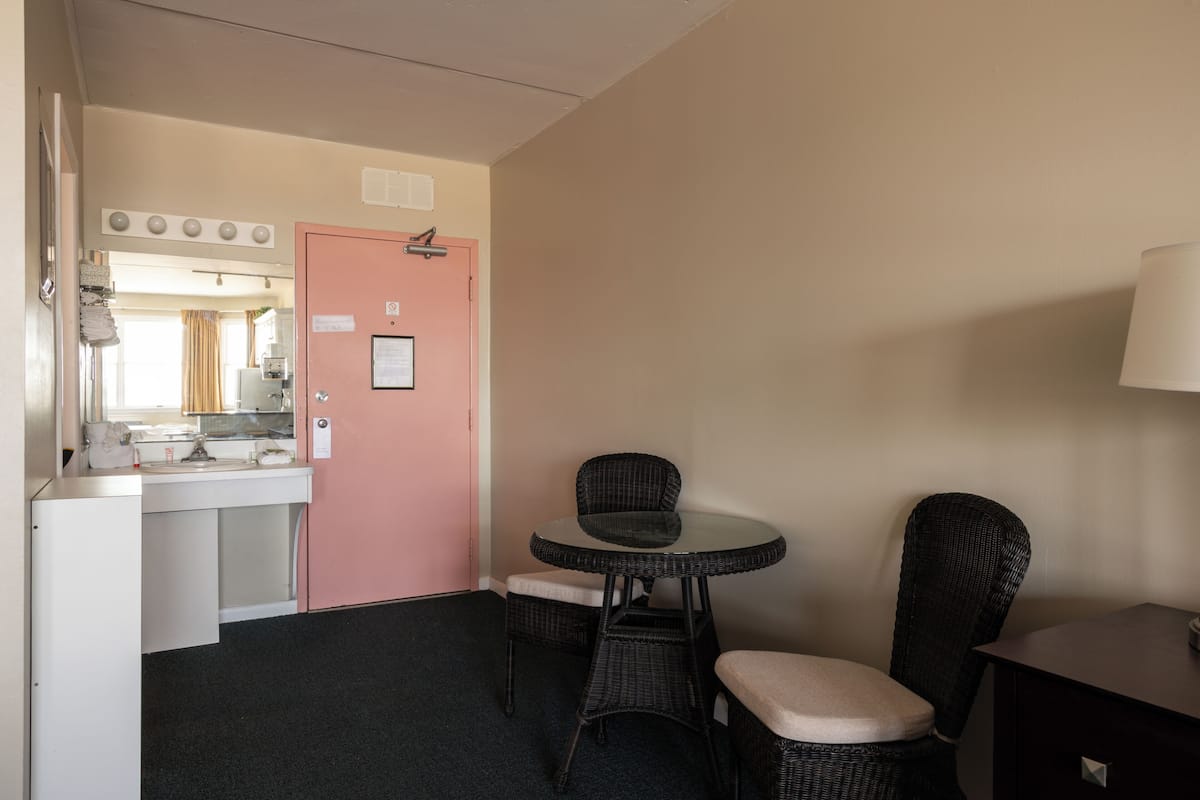 A small room with a pink door, a round table with two chairs, a dark-colored cabinet, and a lamp on a bedside table.