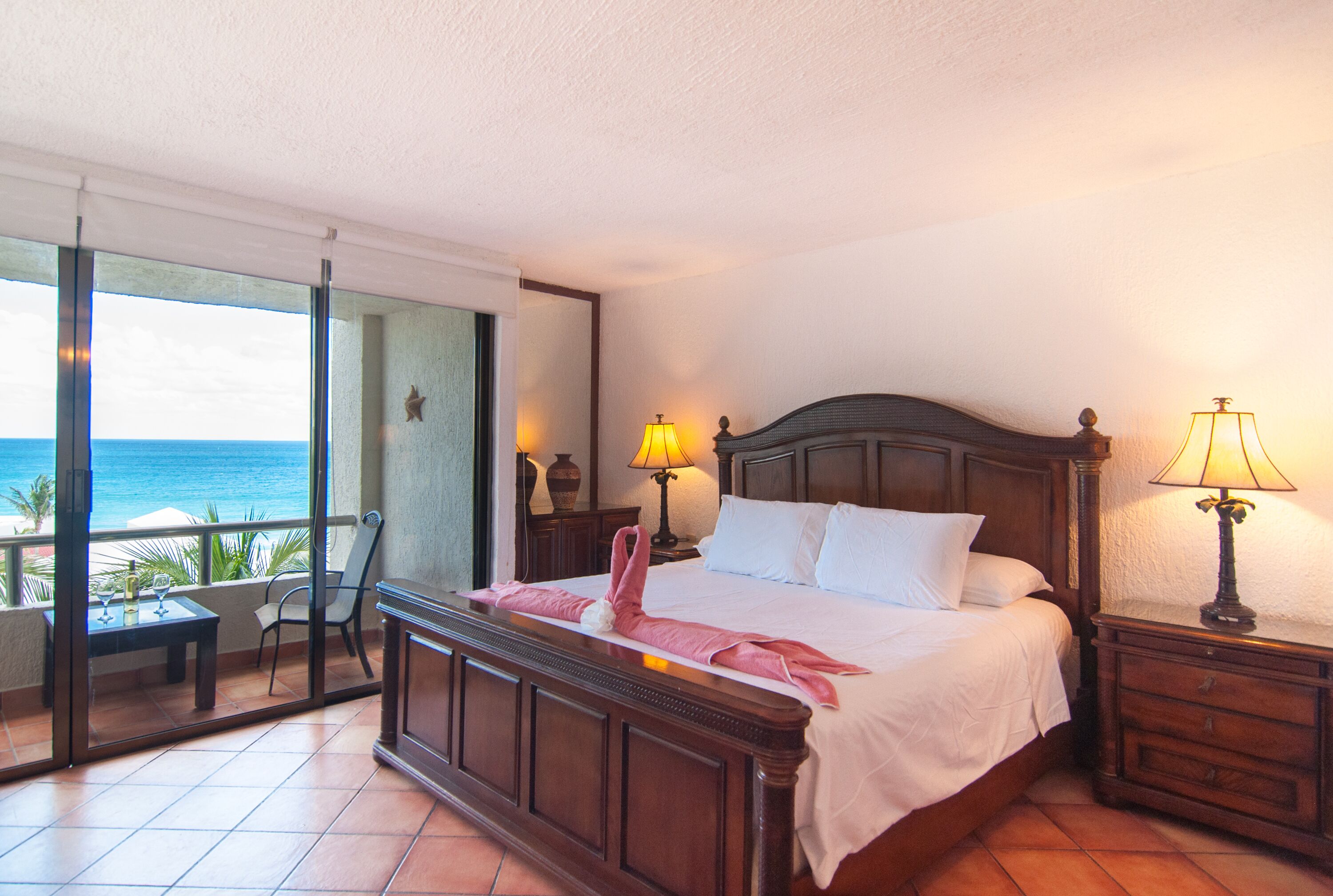Habitación de hotel con una cama grande, dos lámparas de noche, una mesita de noche, un cabecero de madera, una puerta de vidrio que da a un balcón con vistas al océano y una pequeña mesa con sillas.