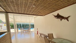 Amplio espacio interior con suelo de baldosas, una mesa de vidrio con sillas, una escultura de pez montada en la pared y vistas a una piscina al aire libre.