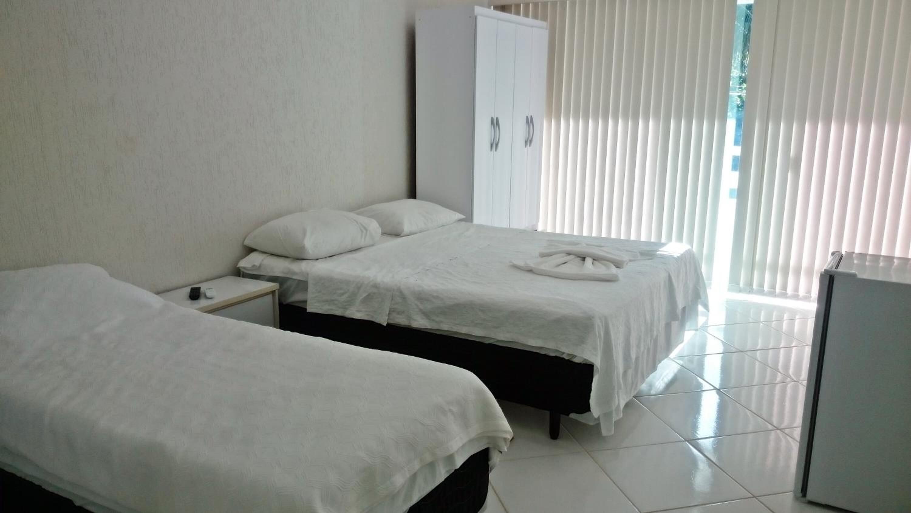 A hotel room with two single beds, a wardrobe, a small table with a remote control, and a refrigerator.