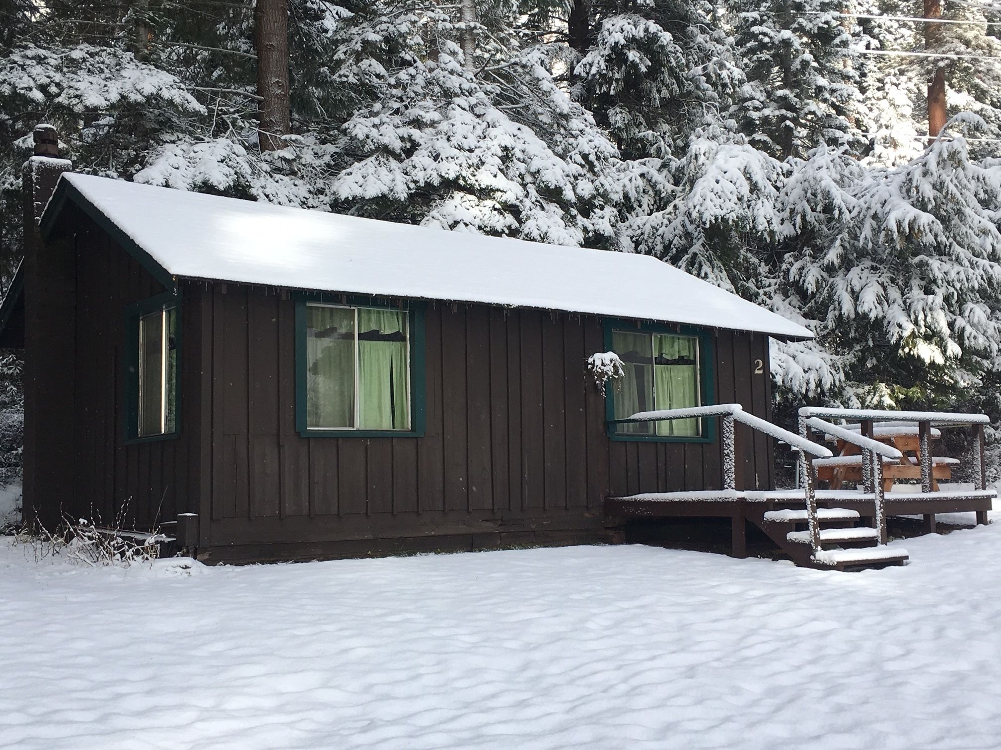 A small cabin with a porch and steps, surrounded by snow-covered trees.