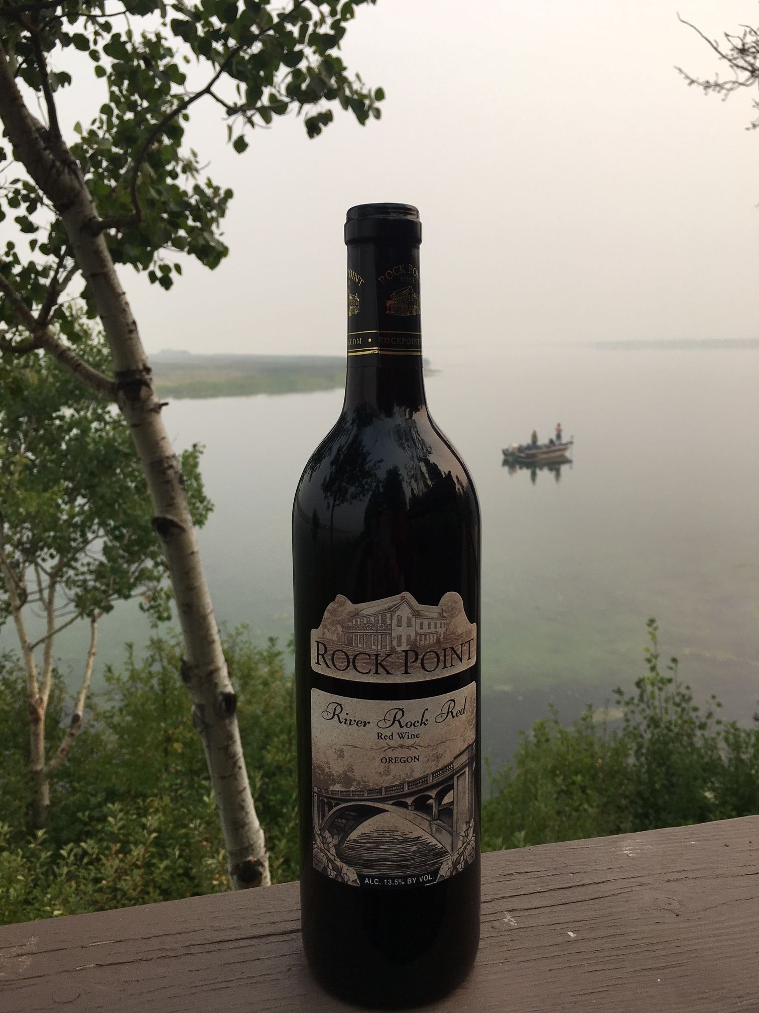 A bottle of River Rock Red wine from Oregon on a wooden surface with a lake and trees in the background.
