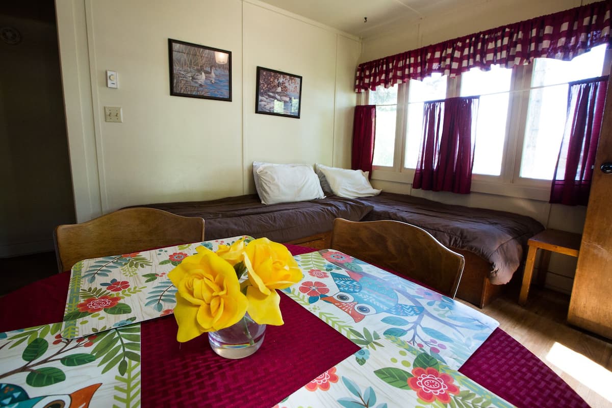 A room with a bed, a table with a floral tablecloth, and two framed pictures on the wall.