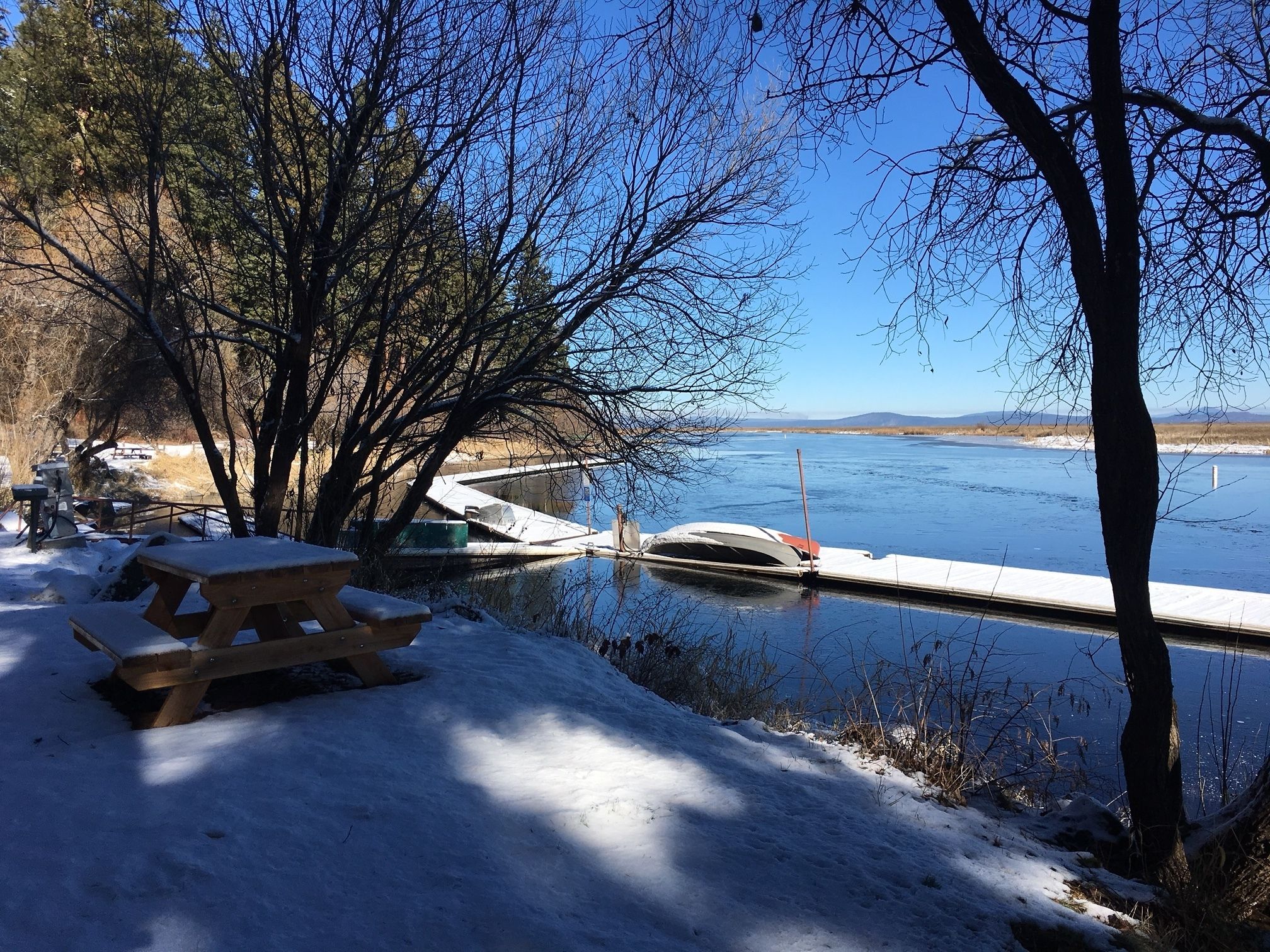 Ett picknickbord och några bänkar står på en snötäckt mark nära en lugn sjö med en brygga.