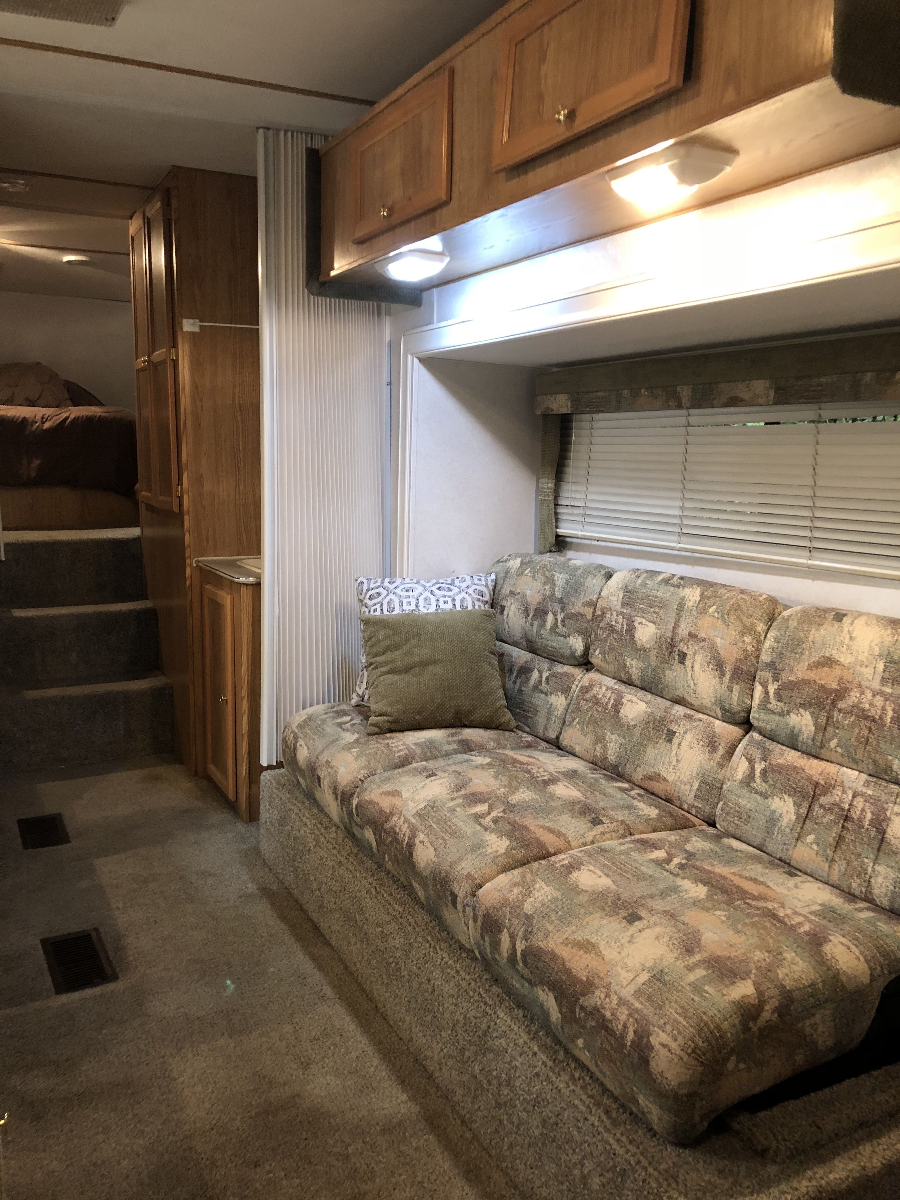 A compact RV interior with a sofa, wooden cabinets, a window with blinds, and a staircase leading to a bed.