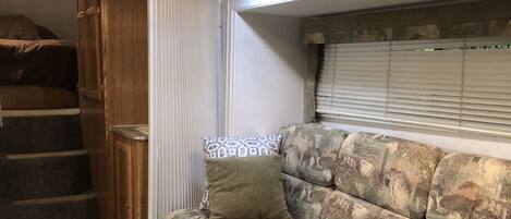 A compact RV interior with a sofa, wooden cabinets, a window with blinds, and a staircase leading to a bed.