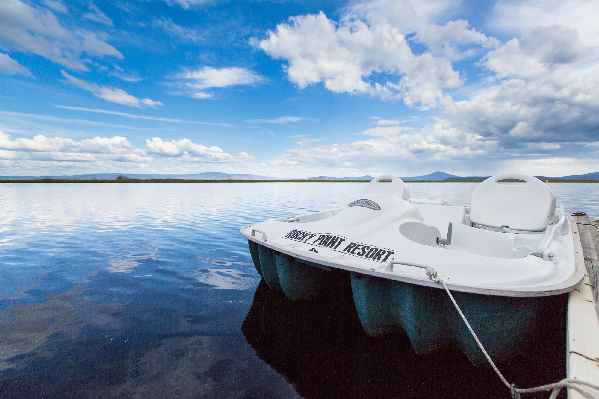 A small white boat with "ROCKY POINT RESORT" written on the side, docked at a calm body of water with a clear sky and distant mountains.