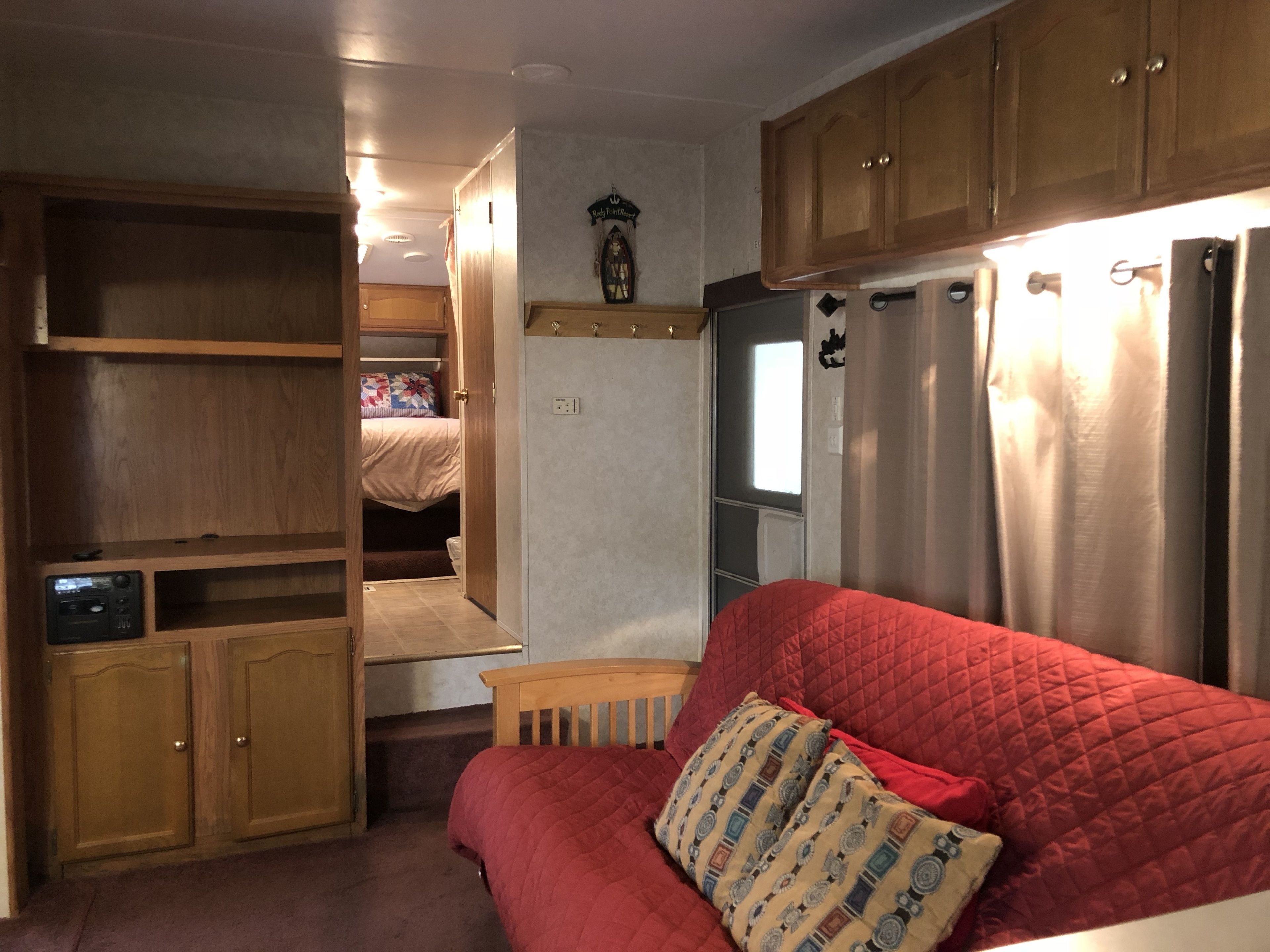 Interior compacto de una autocaravana con kitchenette, una cama con cubierta roja acolchada y armarios de madera.