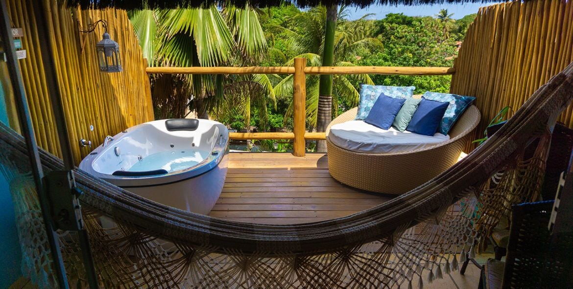 Una hamaca sobre una terraza de madera con un jacuzzi y un sofá de mimbre con cojines.