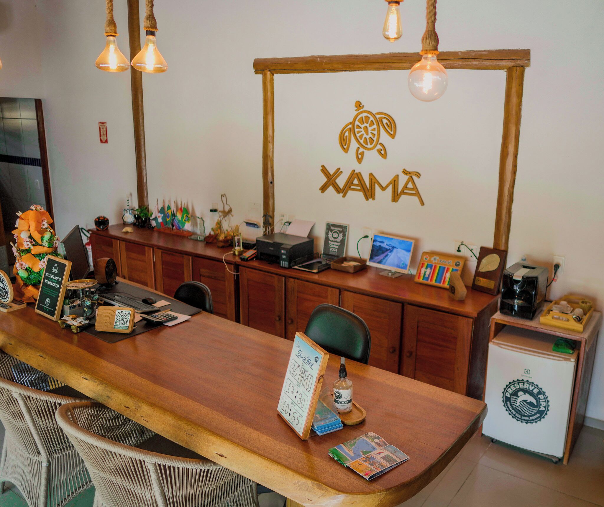 A reception desk with a wooden counter, a sign reading "XAMÁ", and various items like a computer, printer, and decorative objects.