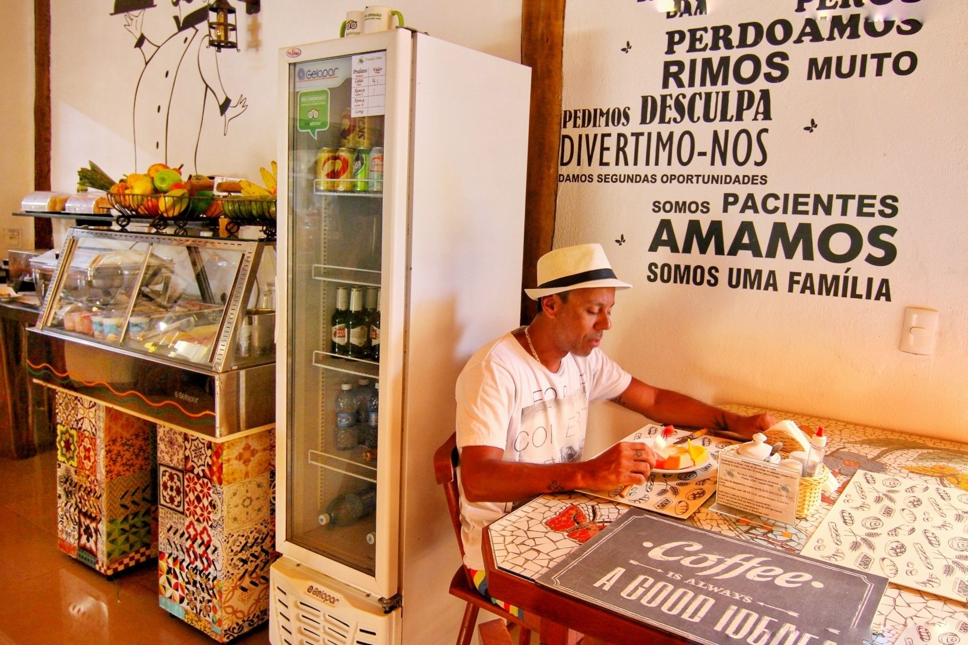 Un hombre sentado a una mesa con un menú de café, un refrigerador lleno de bebidas y una pared con un mensaje en portugués.