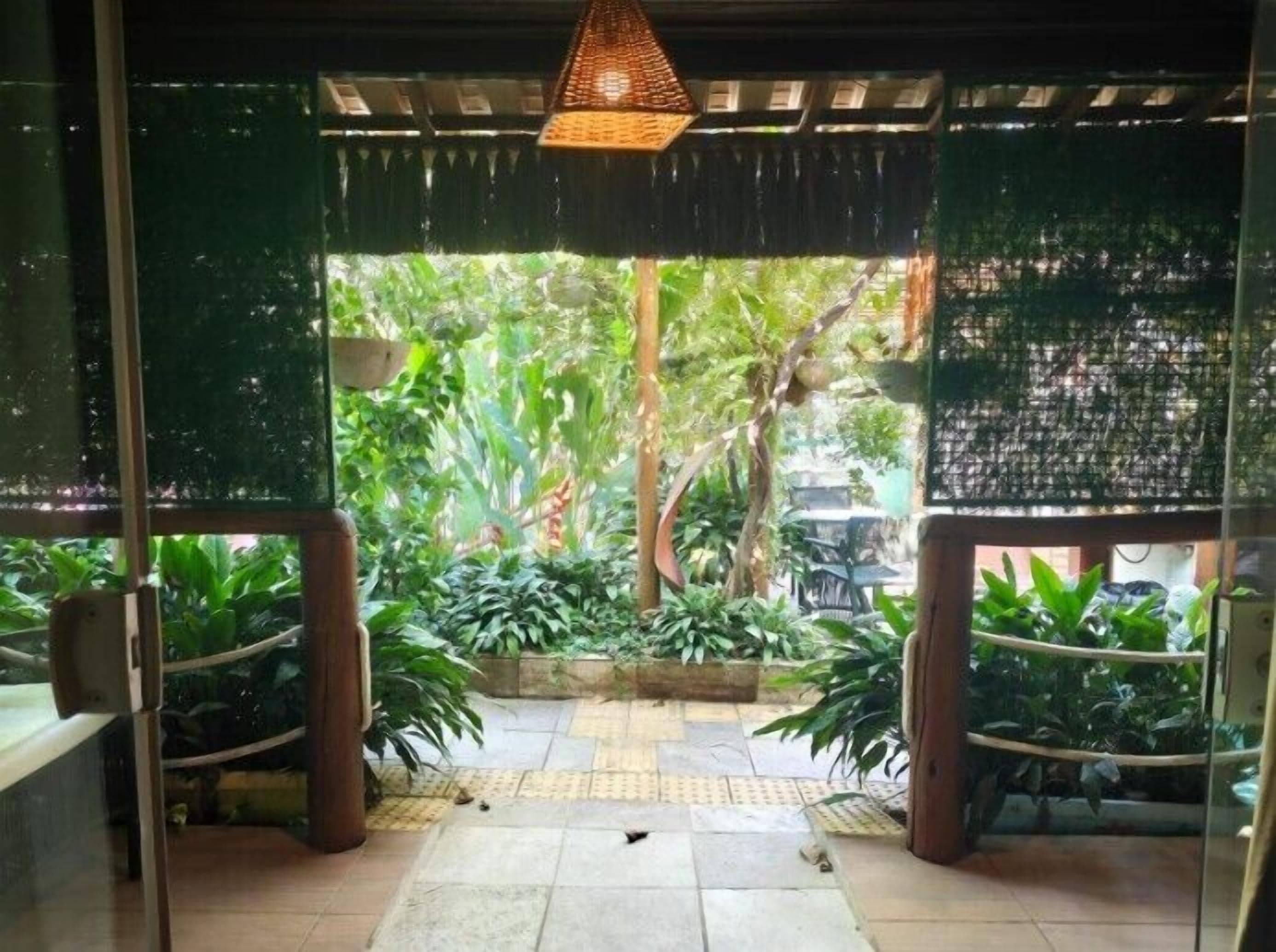 Une entrée avec une porte en verre et un luminaire décoratif en forme de treillis au plafond, qui donne sur un espace jardin agrémenté de plantes en pot et doté d’un sol carrelé.