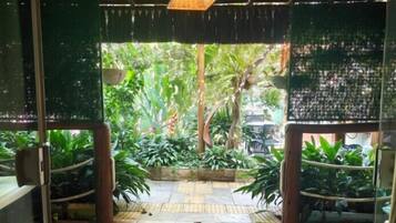 Une entrée avec une porte en verre et un luminaire décoratif en forme de treillis au plafond, qui donne sur un espace jardin agrémenté de plantes en pot et doté d’un sol carrelé.