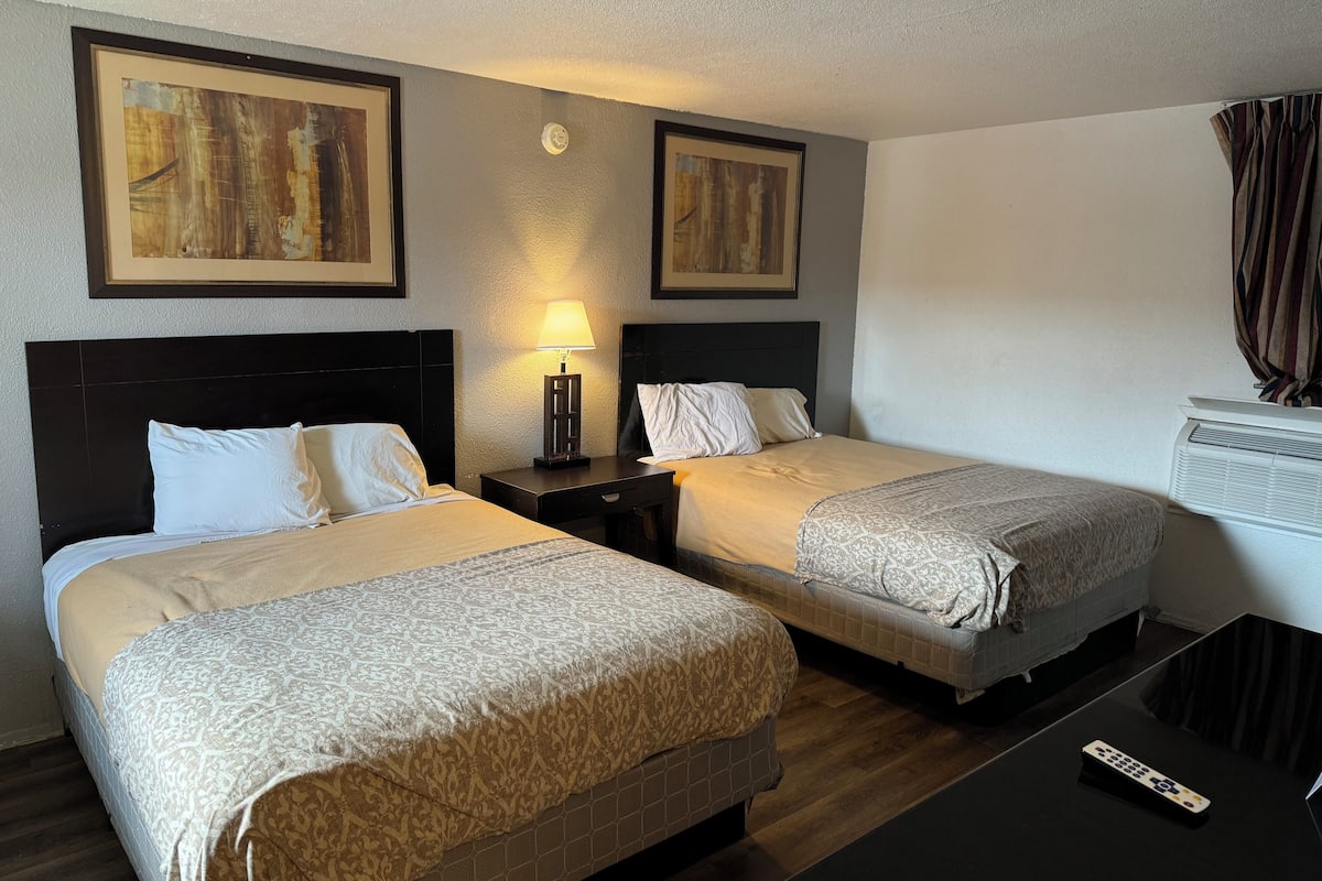 Quarto de hotel com duas camas, uma mesa de cabeceira com luminária, televisão e obras de arte nas paredes.