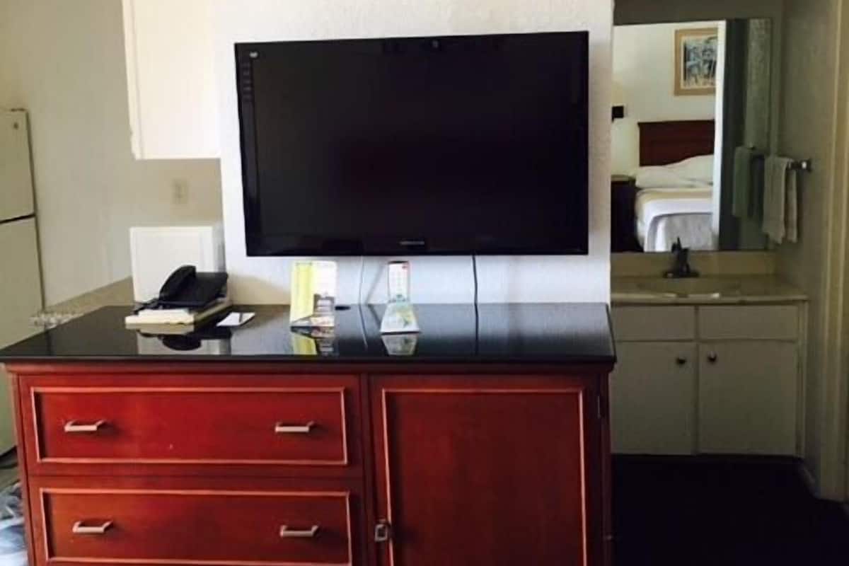 Quarto de hotel com televisão de tela plana fixada em um armário de madeira, cama com colcha estampada branca e pequena área de cozinha compacta com micro-ondas e pia.
