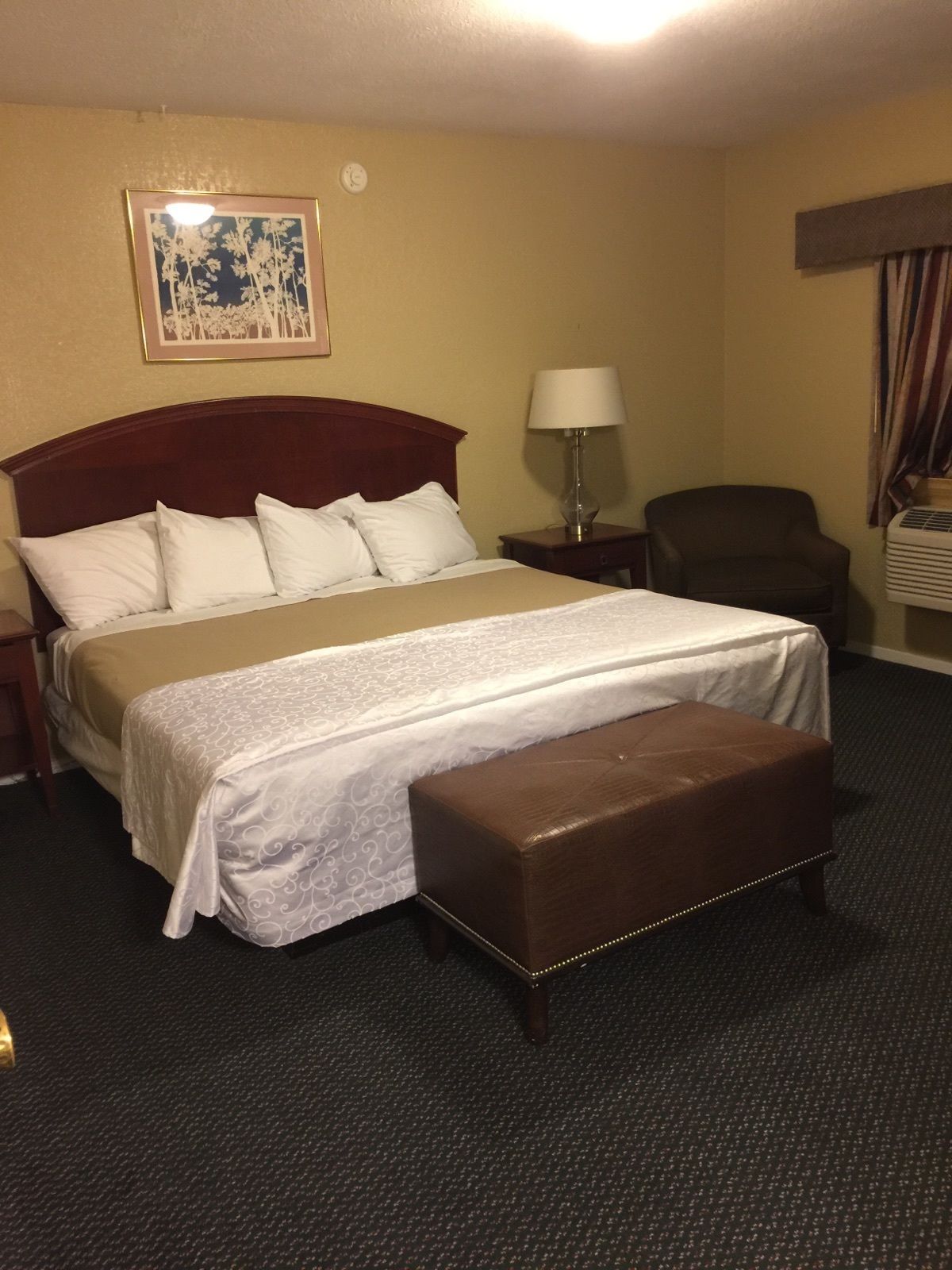 A hotel room with a large bed, a bedside table with a lamp, a chair, and a small table with a basket.