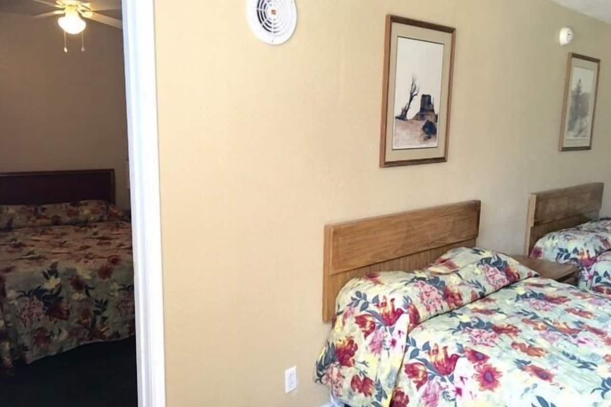 Quarto de hotel com duas camas, roupa de cama floral, um ventilador de teto e duas molduras na parede.