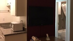 A compact hotel room with a kitchenette, a flat-screen TV mounted on the wall, a wooden dresser, and a view of the bathroom. - Ranch House Inn and Suites (Temple)