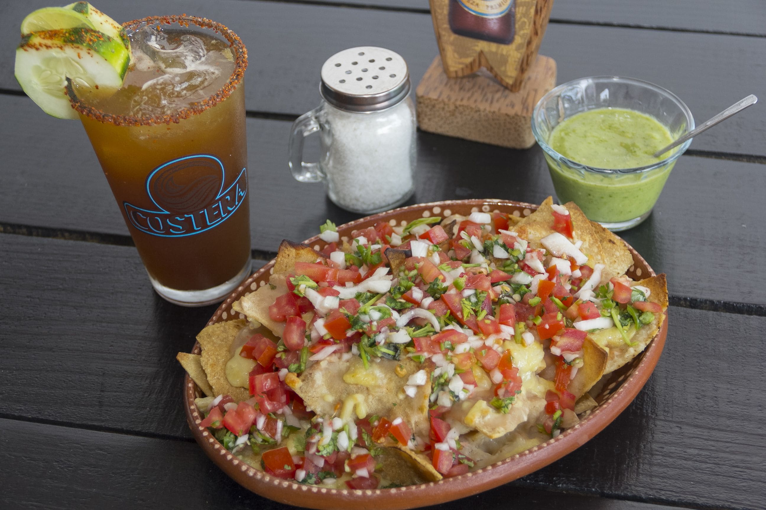 Une assiette de nachos garnie de tomates coupées en dés, d’oignons et de fromage, un verre de thé glacé avec une tranche de citron vert, et un petit bol de sauce verte sur une table en bois.